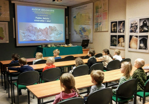 Grupa dzieci przedszkolnych siedź w auli. Z wielkim zainteresowaniem słuchają wykładu na temat: Typów skał, skamieniałości oraz czynników wpływających na rzeźbę terenu.